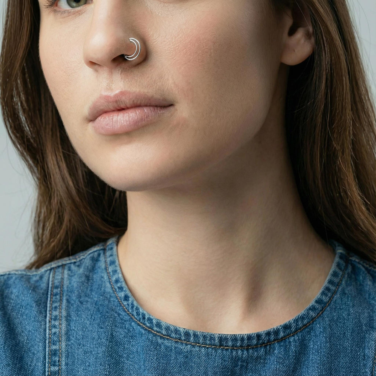 A close-up of a woman's face, focusing on her nose and chin. She is wearing a denim shirt and has a nose ring.