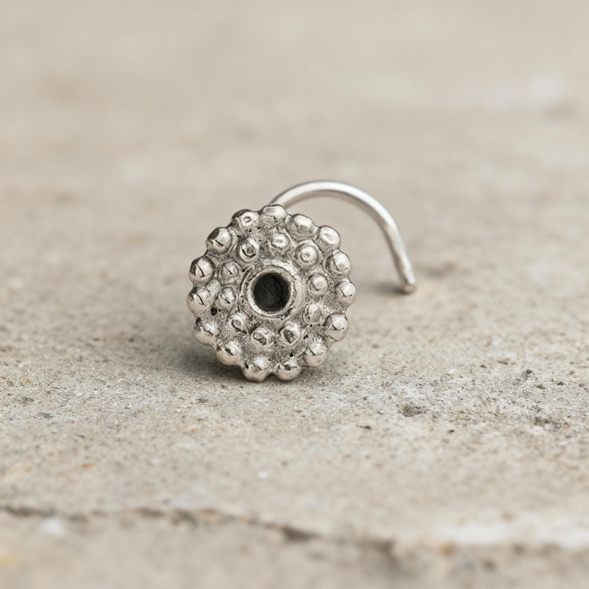 A silver-colored metal nose ring with a circular design and a small hole in the center is shown against a light-colored background.