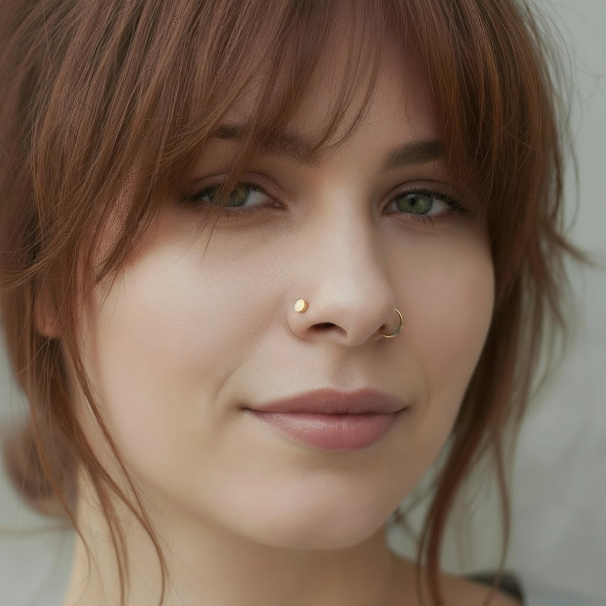 A close-up portrait of a woman with brown hair and a nose piercing, looking directly at the camera with a neutral expression.