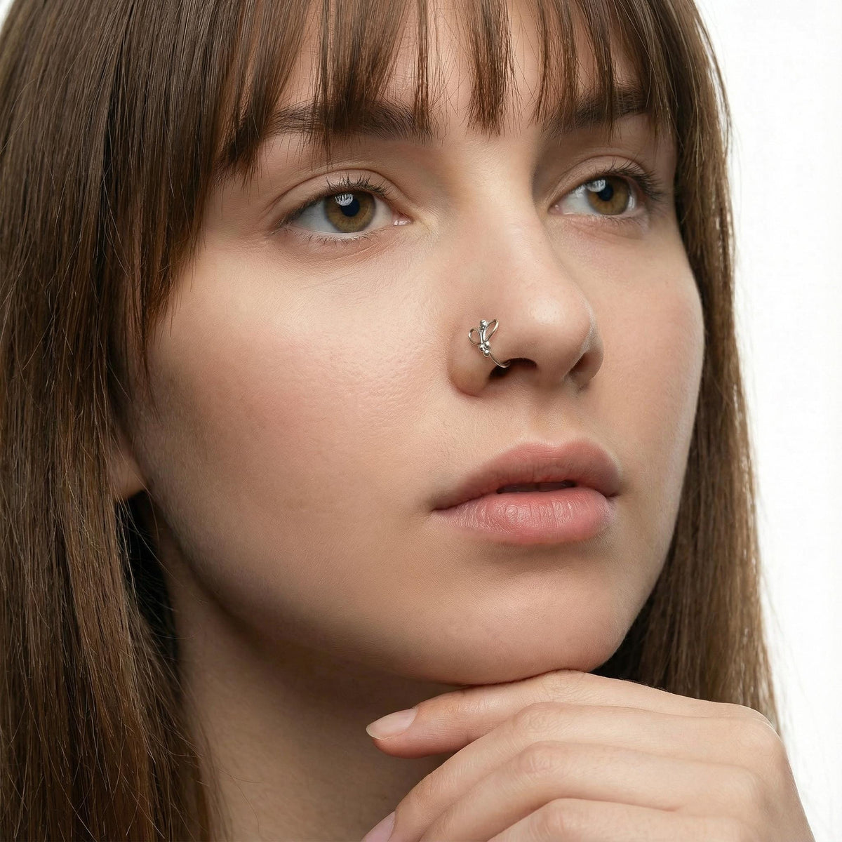 A close-up portrait of a young woman with long brown hair, wearing a nose ring, looking pensive with her chin resting on her hand.