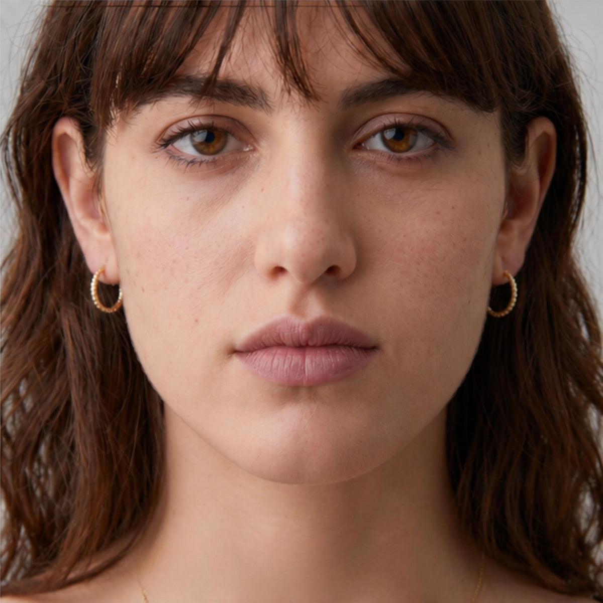 A close-up portrait of a woman with brown hair and gold hoop earrings, looking directly at the camera with a neutral expression.