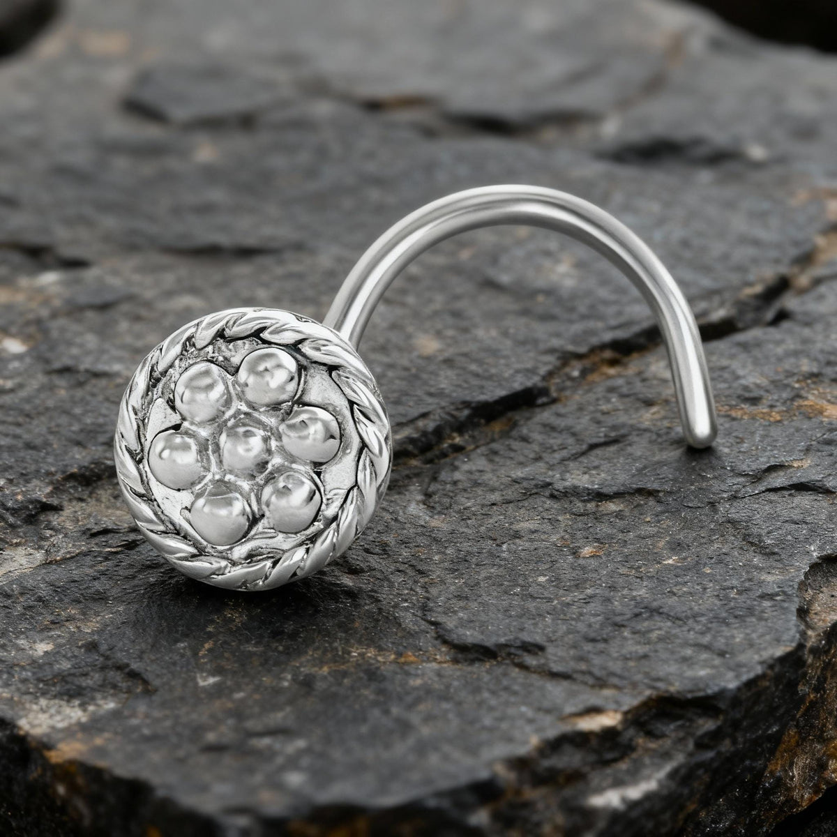 A silver nose ring with a circular design and a curved metal piece is resting on a dark, rocky surface.