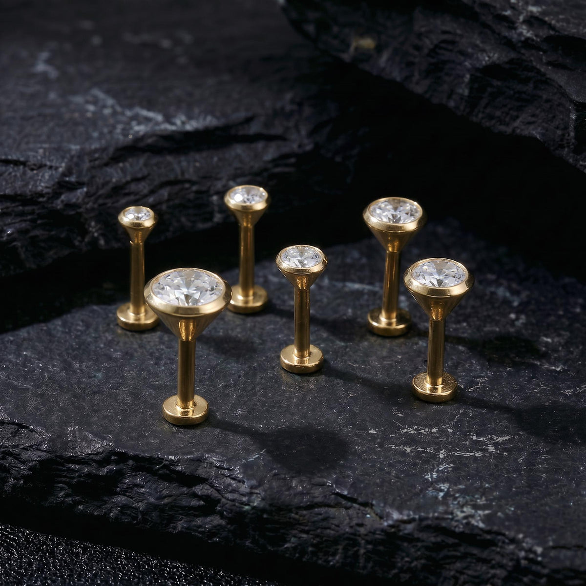 A group of six gold-colored diamond-encrusted earrings or studs is arranged on a dark, textured surface, such as a stone or a black background.
