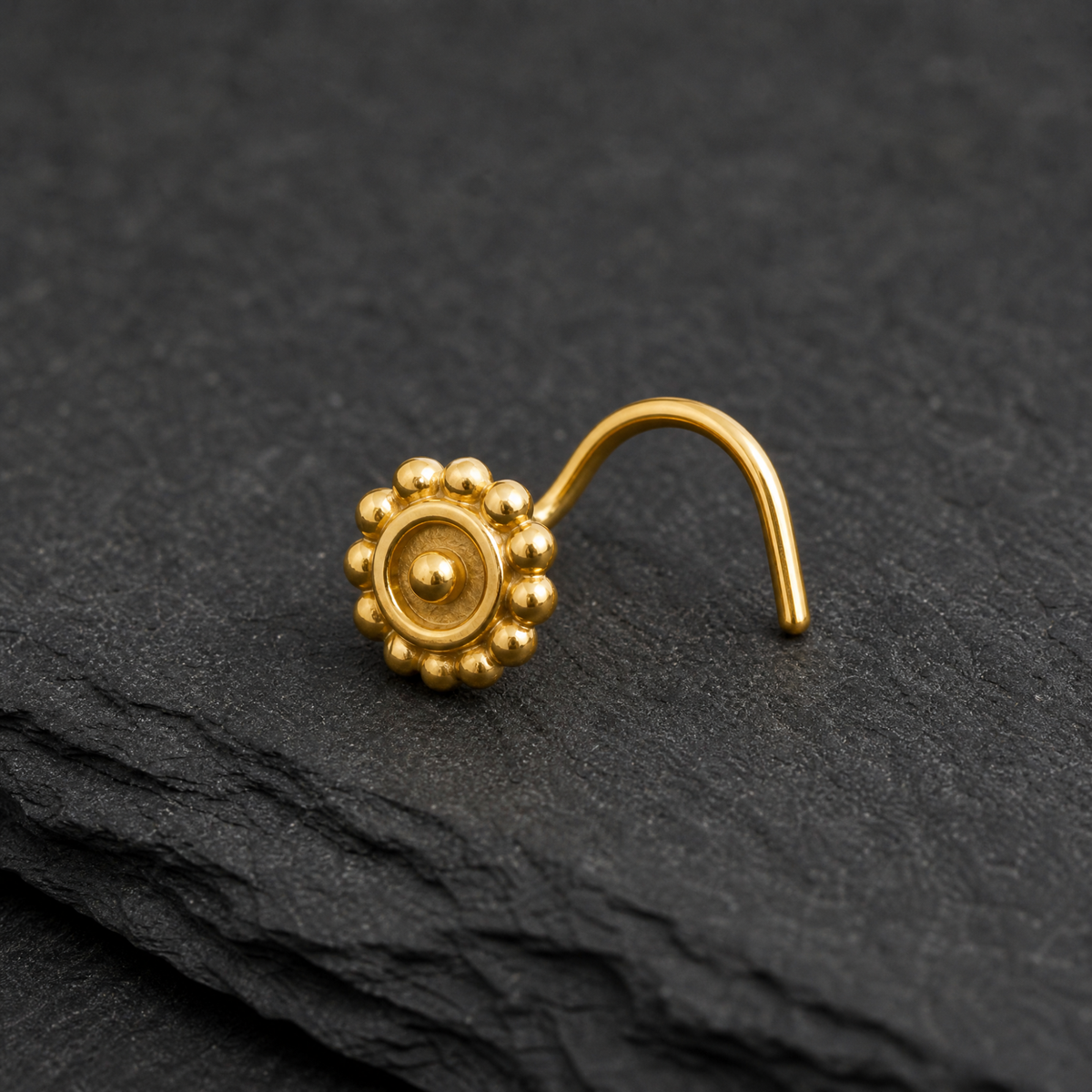 A gold-colored nose stud with a decorative flower-like design is resting on a dark gray surface.