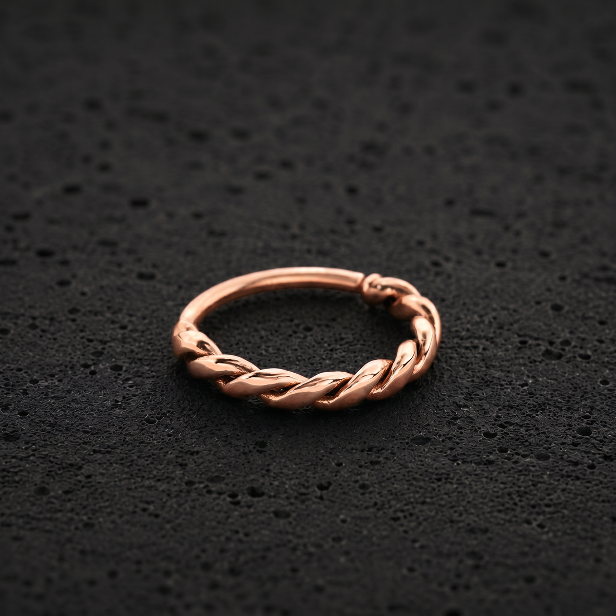 A copper-colored ring with a twisted, braided design is shown against a dark, textured background.