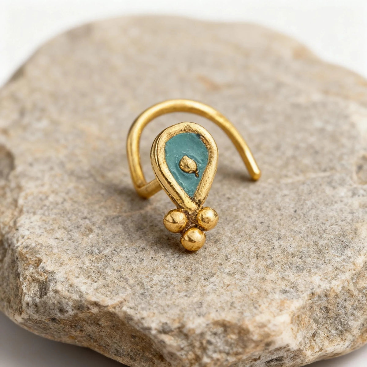 A gold-colored nose ring with a turquoise stone in the center, resting on a gray rock.