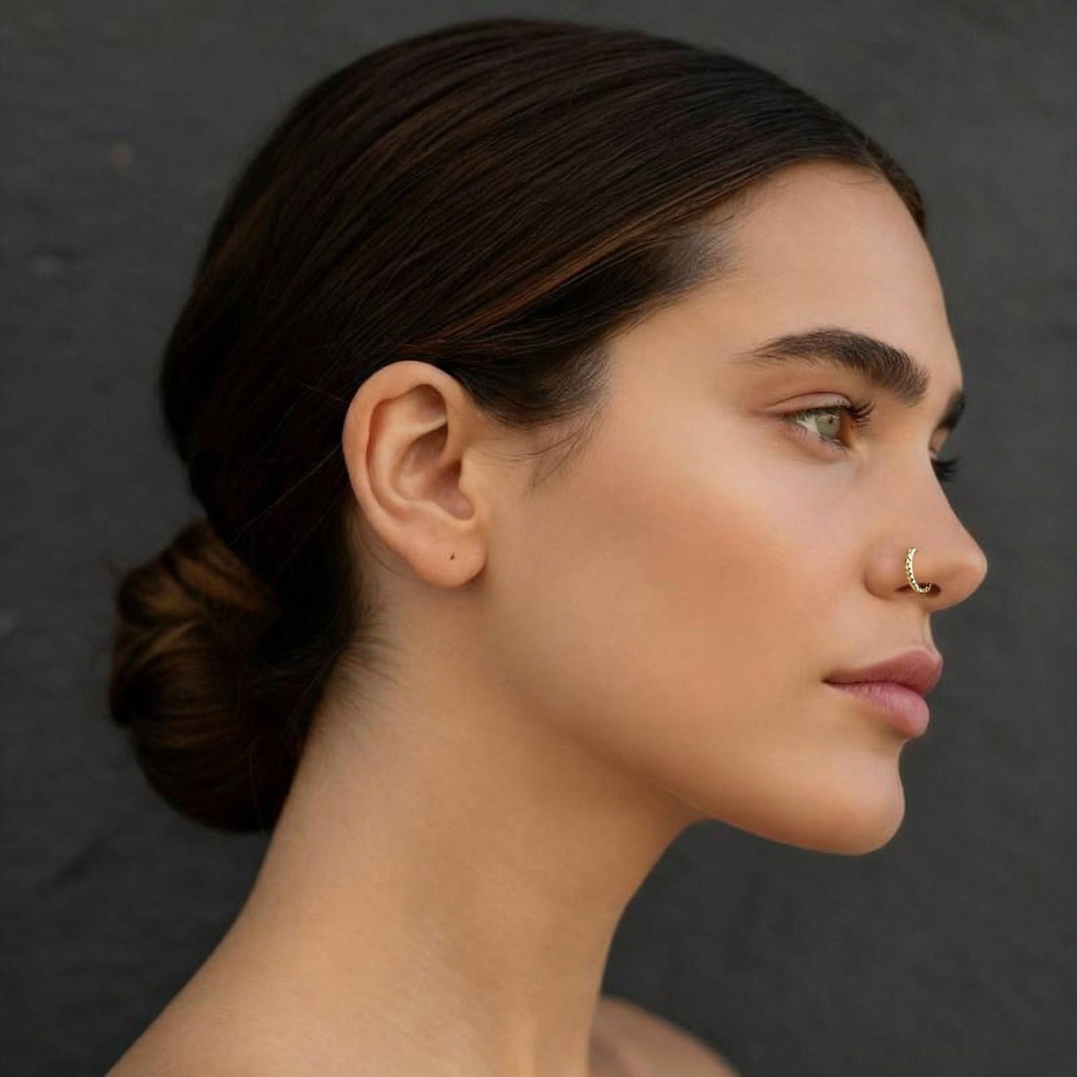 A close-up profile view of a woman with dark hair and a nose ring, looking to the side against a dark background.