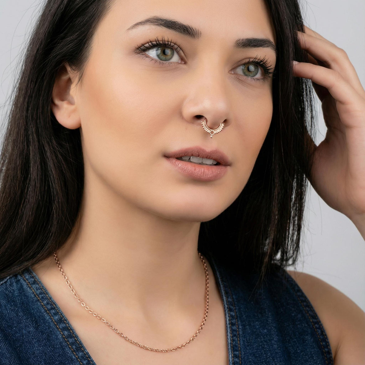 A close-up portrait of a woman with long dark hair, wearing a necklace and a nose ring. She is looking off to the side with a thoughtful expression.