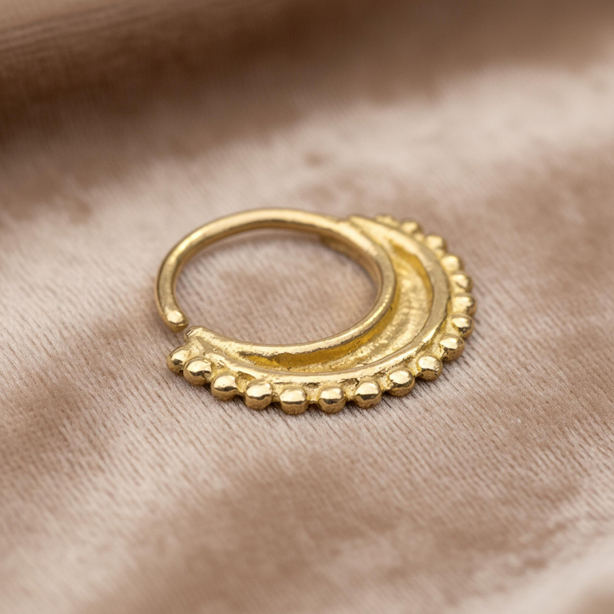 A gold-colored metal nose ring with a decorative design is shown against a textured beige background.