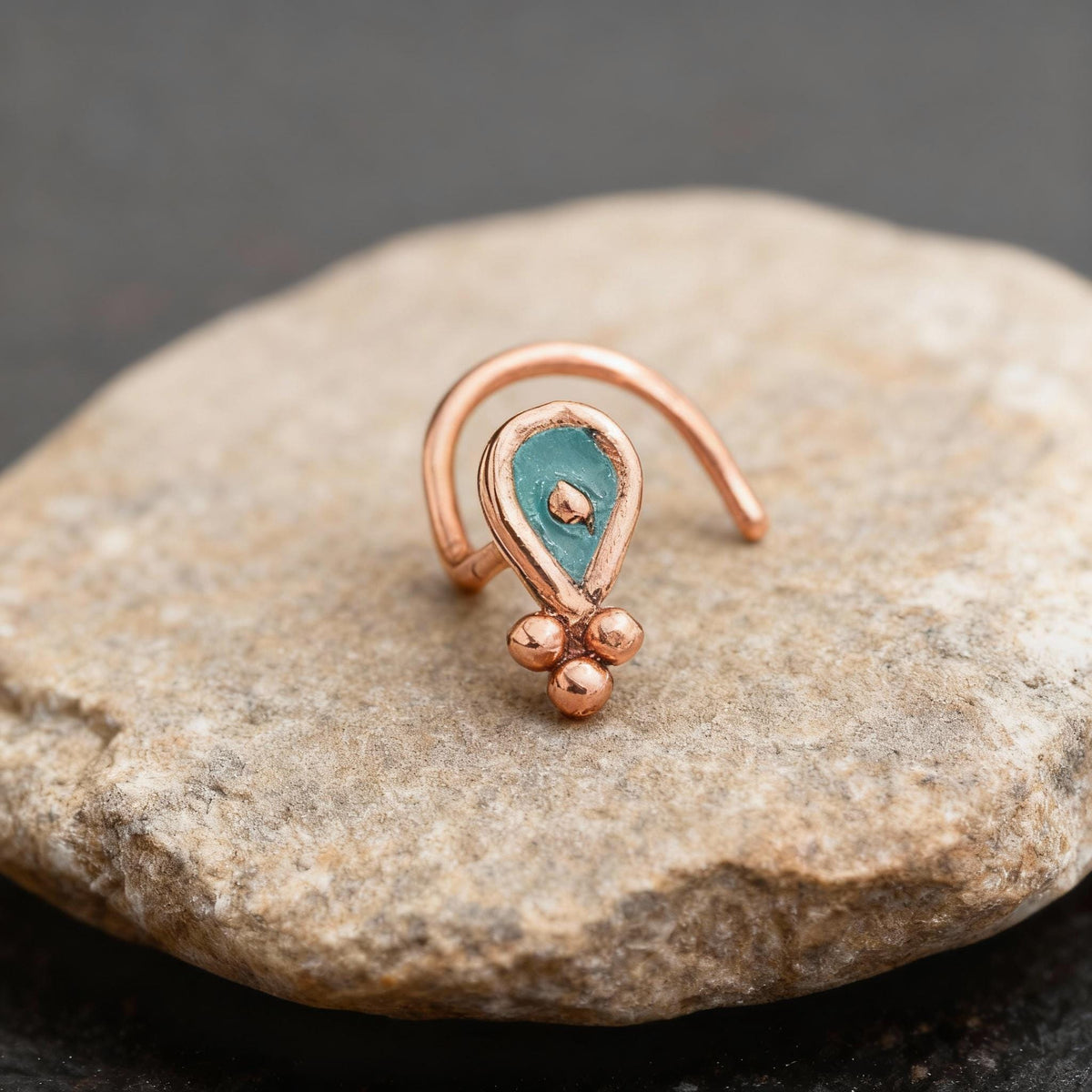 A copper-colored nose ring with a teal-colored stone in the center, resting on a rock.