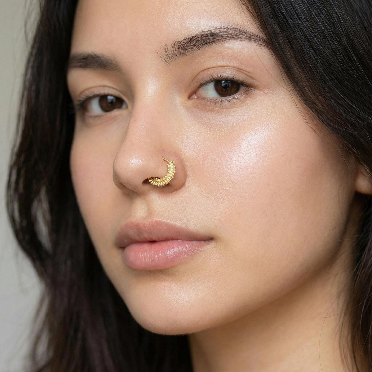 A close-up portrait of a young woman with dark hair, wearing a gold-colored nose ring.
