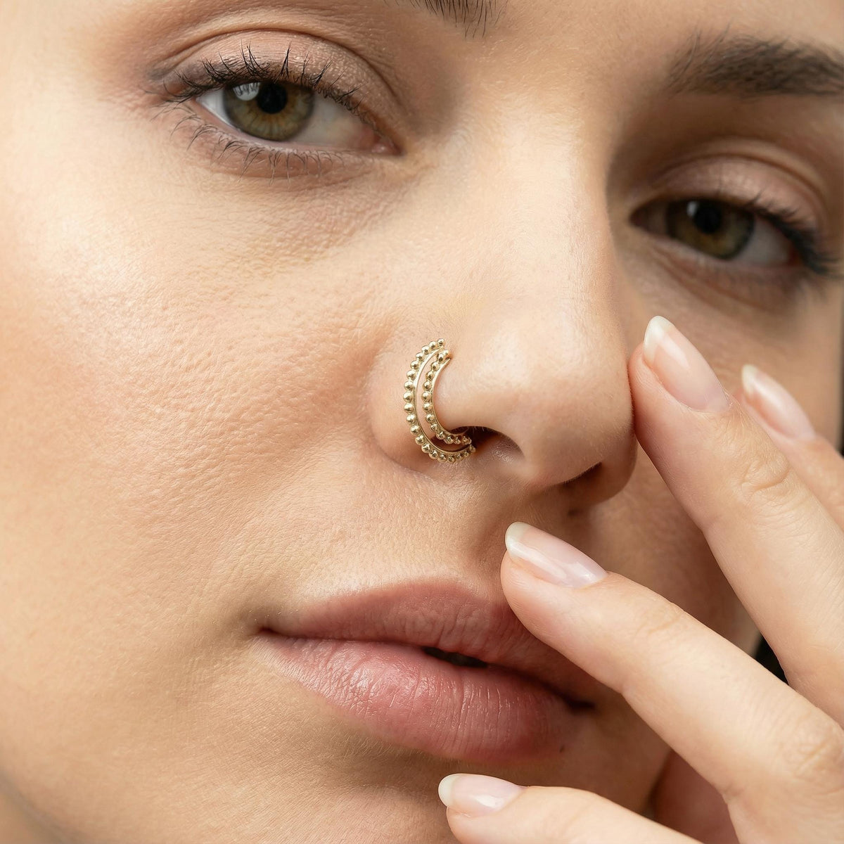 A close-up of a woman's face, focusing on her nose and fingers. She is wearing a gold nose ring.