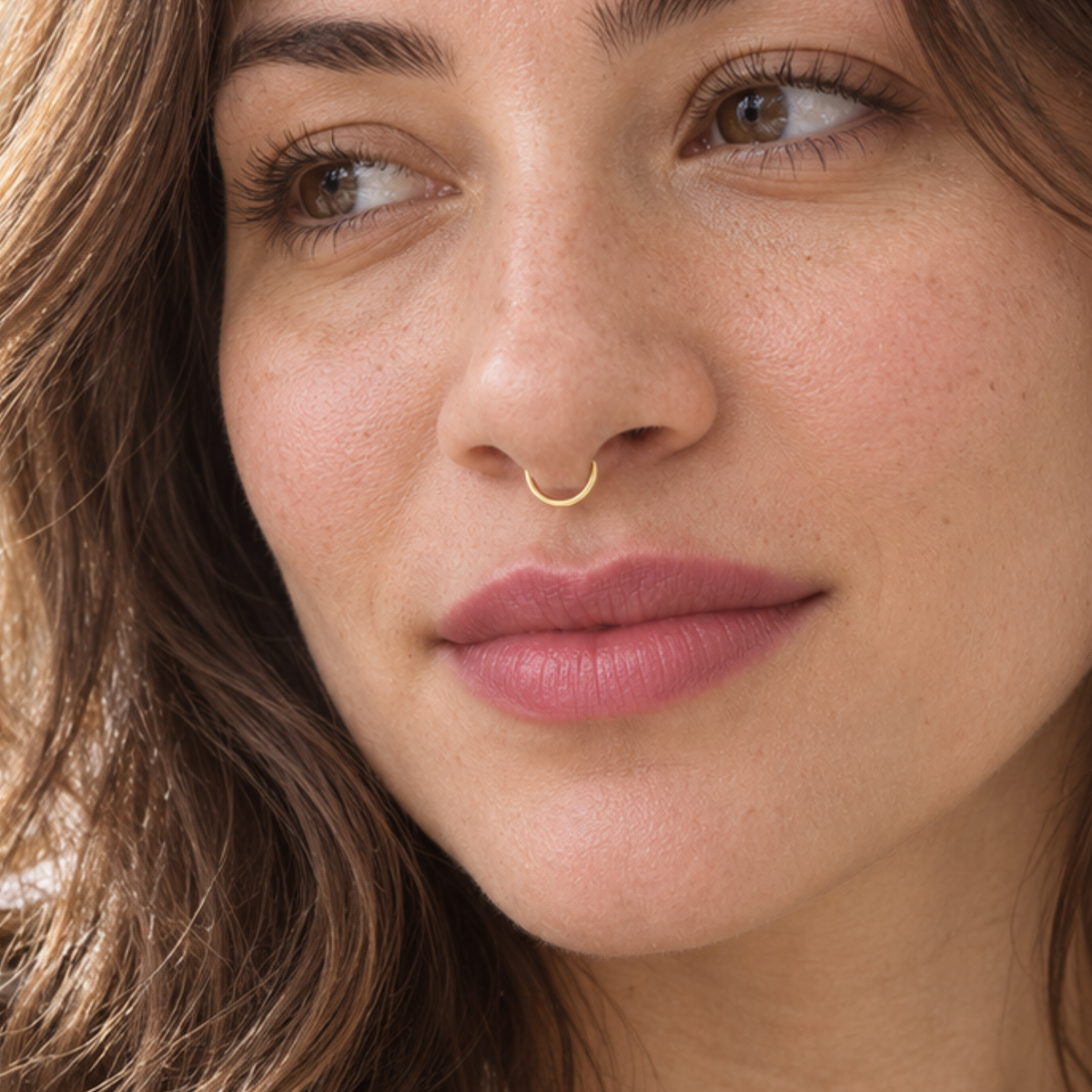 A close-up portrait of a woman with long, wavy brown hair and a gold nose ring.