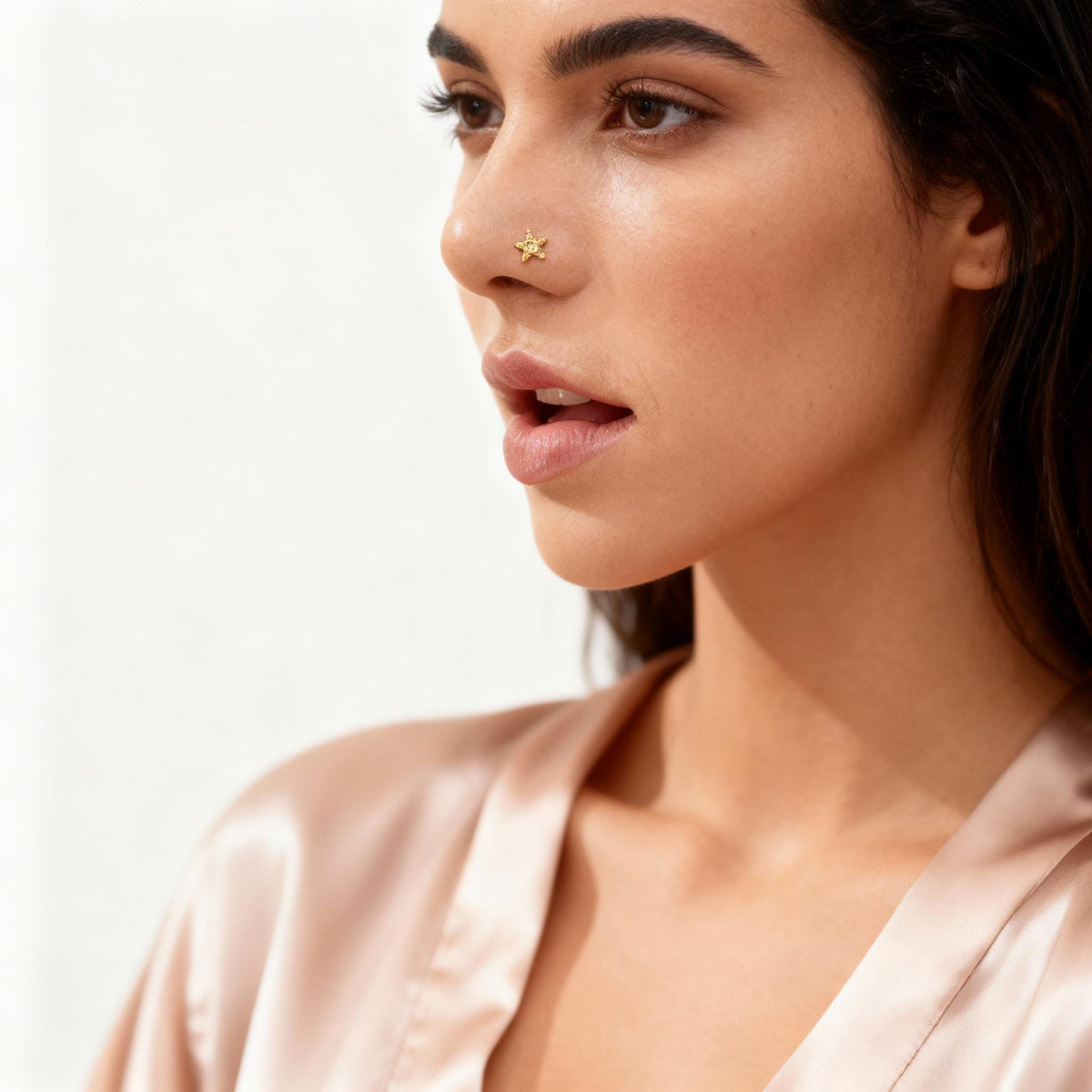 A close-up portrait of a woman with dark hair, wearing a pink satin robe, and a nose stud.