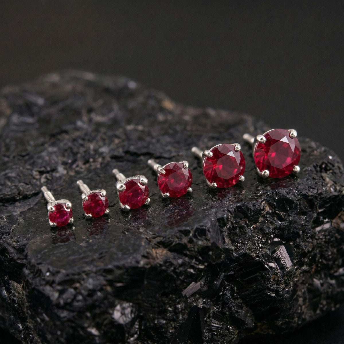 A collection of red gemstone earrings in various sizes, ranging from small to large, resting on a black, rocky surface.