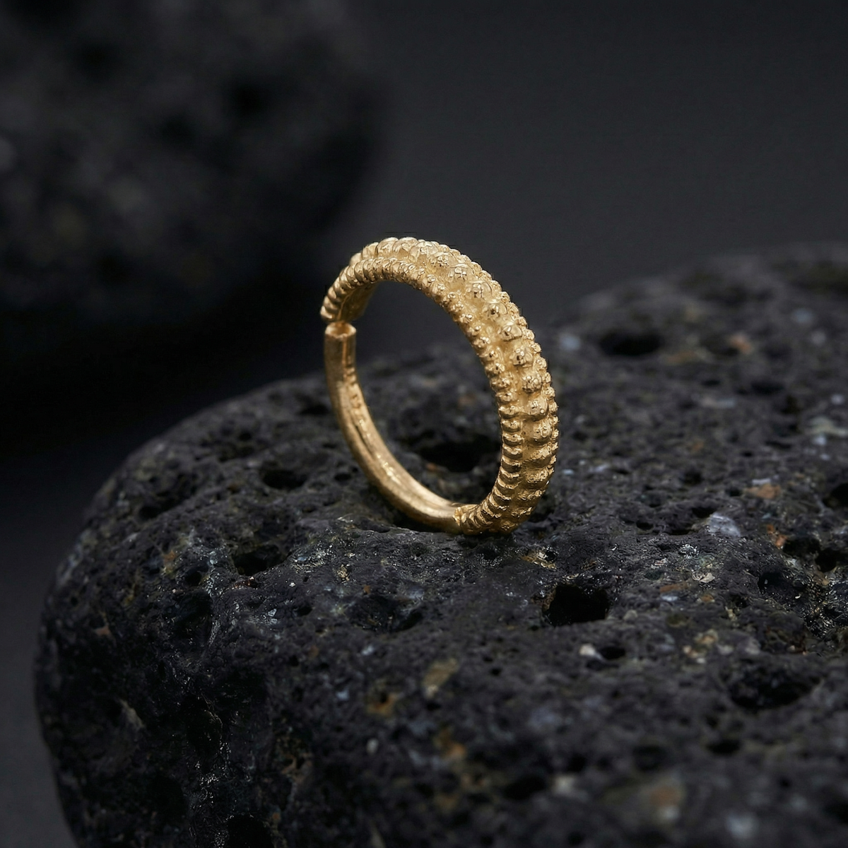 A gold ring with a textured design sits on a dark, rocky surface.