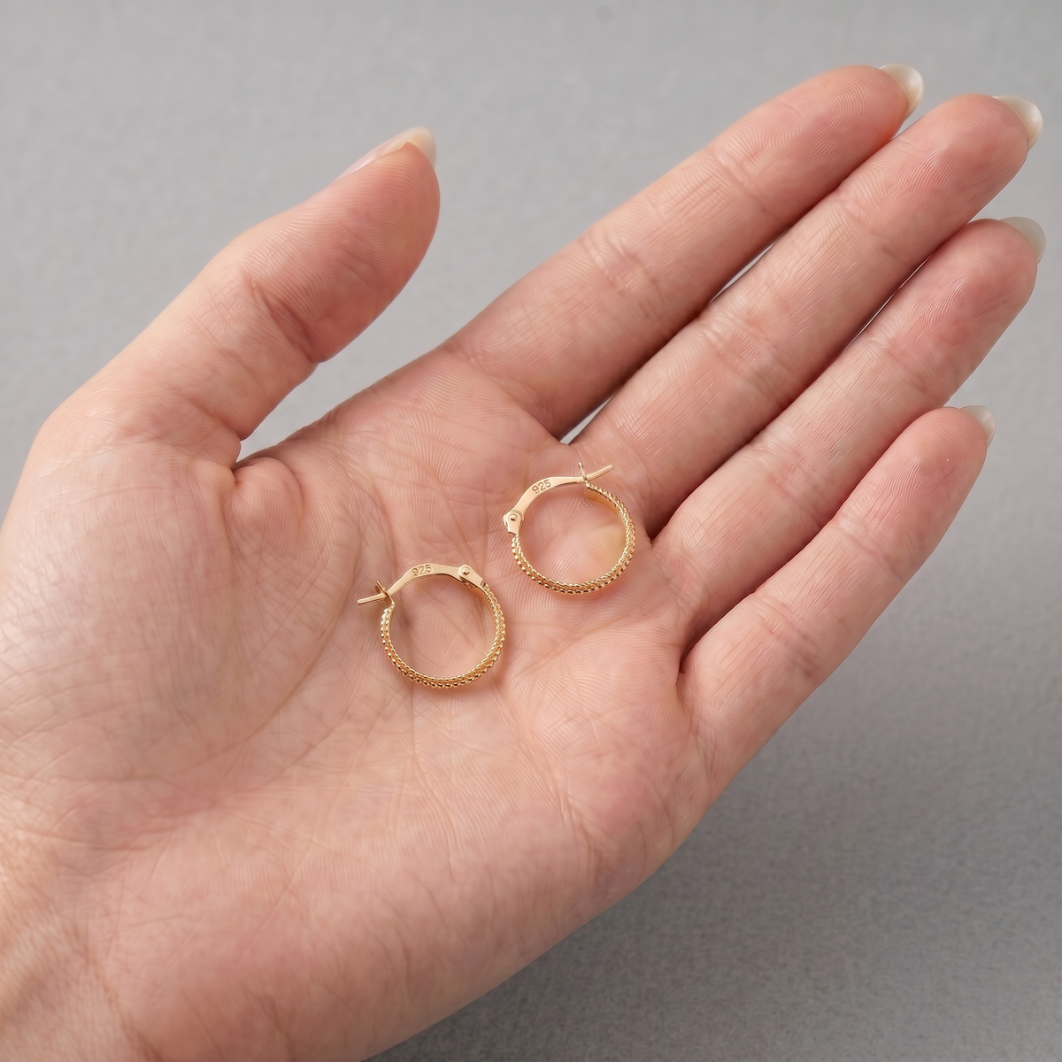 A hand holding a pair of gold hoop earrings against a gray background.