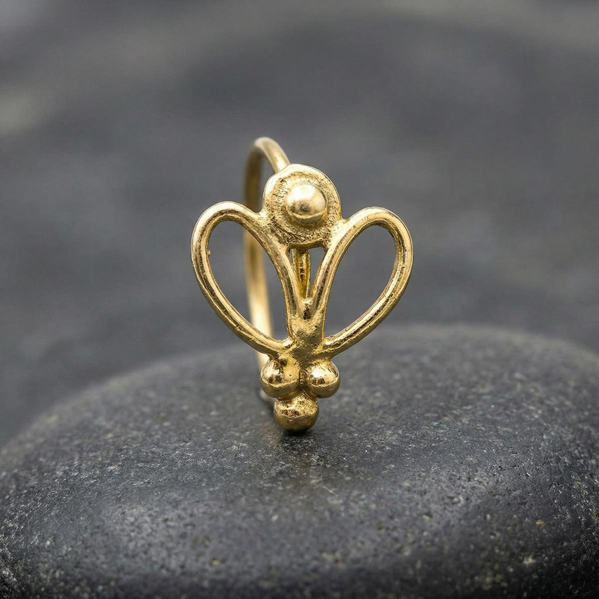 A gold-colored metal nose ring with a floral design is shown against a dark background.