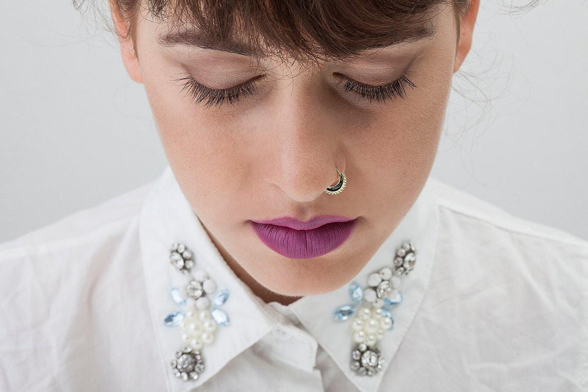 A close-up portrait of a woman with her eyes closed, wearing a white collared shirt with a necklace of rhinestones and a nose ring.