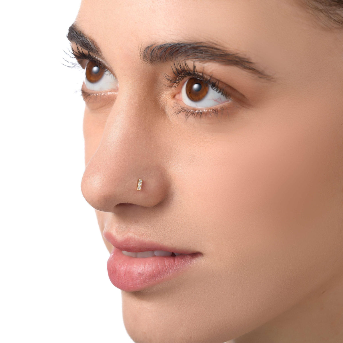 A close-up portrait of a woman with brown eyes and a nose stud, looking to the side with a thoughtful expression.